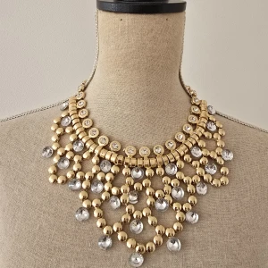 Statement halsband i guld med kristaller - Ett riktigt maffigt statement halsband i guldton med stora klara kristaller och glittriga detaljer. Halsbandet har flera rader av guldfärgade kulor och gnistrande stenar som ger en lyxig och iögonfallande look. Perfekt för dig som vill sticka ut och glänsa.