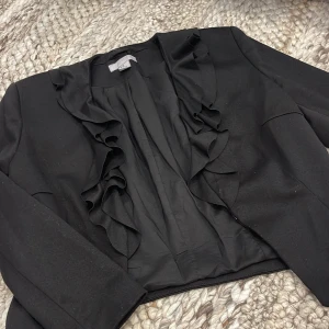 Svart kavaj med volang från H&M - Svart kavaj från H&M i storlek 40 med snygga volangdetaljer längs framkanten. Klassisk modell med lång ärm och croppad passform. 
