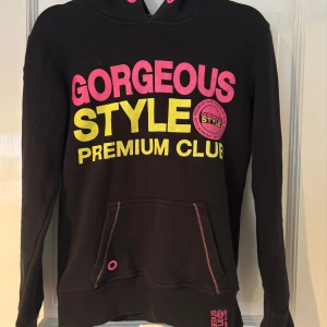 Svart hoodie från Fishbone med tryck - Svart hoodie från Fishbone med stora tryck i rosa och gult framtill. Har huva med rosa öljetter, magficka och ett FS-märke med nitar på ärmen. Perfekt för dig som gillar streetstyle och färgstarka detaljer.