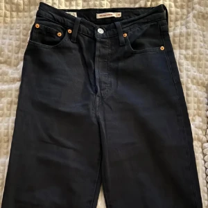 Svarta Levi's Ribcage Ancle jeans straight fit - Klassiska svarta Levi's Ribcage Ancle jeans med raka ben och hög midja. Jeansen har fem fickor, kopparfärgade nitar och den ikoniska läderpatchen bak. Tillverkade i robust denim som ger en tidlös look. Perfekta för dig som gillar en clean och stilren vibe.