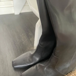 Svarta höga stövlar i skinn - Stilrena svarta stövlar med fyrkantig tå och högt skaft. Tillverkade i slätt skinn med minimalistisk design och platt sula. Perfekta för dig som gillar en clean och edgy look till höst och vinter.