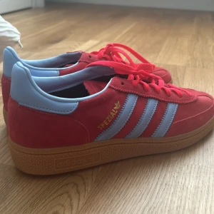 Adidas Spezial röda sneakers med blå detaljer  - Snygga Adidas Spezial sneakers i rött mocka med ljusblå detaljer och klassiska tre ränder på sidorna. Skorna har gummisula, snörning och guldfärgad Spezial-logga. Perfekta för dig som vill sticka ut med färg och retrostil. Helt nya, Vill inte ha de eftersom att de passar inte så mycket med min stil.