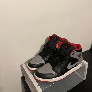 Nike Air Jordan 1 Mid Svart/Grå/Röd - Snygga Nike Air Jordan 1 Mid med svart och grå ovandel i skinn, röda detaljer och röd yttersula. Klassisk Jordan-logga på sidan och röd Jumpman på plösen. Perfekta för dig som vill sticka ut med en ikonisk sneaker.