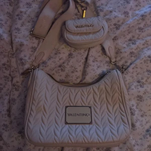 Beige quiltad axelväska Valentino - Säljer en beige quiltad axelväska från Valentino med matchande liten väska på axelbandet. Väskan har ett snyggt mönster, bred axelrem och metallskylt framtill. Perfekt för dig som vill ha en trendig och praktisk väska med plats för det viktigaste.