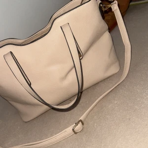 Beige axelväska med dragkedja - Stilren beige axelväska med svarta kanter och guldfärgade detaljer. Väskan har både handtag och justerbar axelrem samt dragkedja upptill och ett extra fack med dragkedja på utsidan. Insidan är rymlig med flera fack för småsaker. Perfekt för dig som vill ha plats för allt.