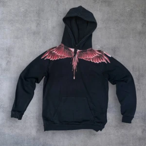 Marcelo Burlon svart hoodie med vingar - Svart hoodie från Marcelo Burlon med ikoniskt vingtryck i rött och rosa över axlar och bröst. Tröjan har huva, känguruficka och ribbade muddar. Grym streetwear-vibe och perfekt för dig som vill sticka ut.