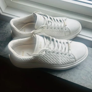 Vita sneakers från BOSS i skinn - Snygga vita sneakers i skinn från BOSS med gulddetaljer och diskret logga på plös och häl. Skorna har en stilren design med mönstrade partier på sidorna och klassisk snörning. Storlek 38. Nypris: 2500:-