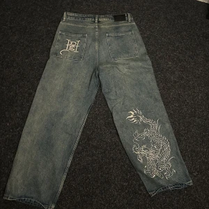 Ed Hardy jeans med drakbrodyr - Unika blå jeans från Ed Hardy med stor vit drakbrodyr på ena benet och broderade loggor fram och bak. Byxorna har raka ben, stora fickor och är tillverkade i klassiskt denim. Perfekta för dig som vill sticka ut med streetwear-vibe. Storlek 30/30