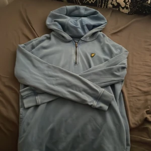 Ljusblå hoodie från Lyle & Scott - Ljusblå hoodie från Lyle & Scott med halv dragkedja och klassisk gul logga på bröstet. Tröjan har huva, långa ärmar och ribbade muddar. Perfekt för en chill och avslappnad stil.