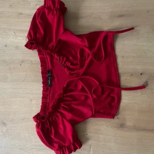 Röd croppad topp från Boohoo - Superfin röd croppad topp från Boohoo med puffiga korta ärmar och volangdetaljer vid axlar och ärmslut. Toppen har offshoulder-modell och knytband framtill. Materialet är ribbat och stretchigt för en skön look.