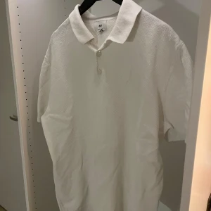 Vit pikétröja från H&M - Klassisk vit pikétröja från H&M i regular fit. Tröjan har korta ärmar, krage och två knappar framtill. Materialet är strukturerat och känns mjukt mot huden, perfekt för en clean och stilren look. Aldrig använd