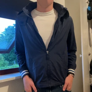 Blå vindjacka Tommy Hilfiger 164 - Snygg marinblå vindjacka från Tommy Hilfiger med huva och dragkedja. Jackan har vita och blå ränder på mudden, diskret logga på ärmen och meshfoder på insidan. Perfekt för dig som gillar sportig och stilren look. Storlek 164 men passar som xs✍🏻