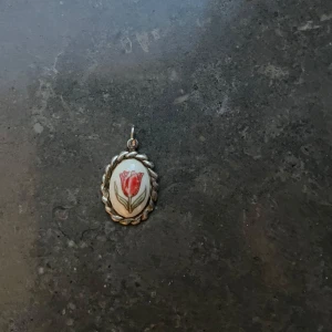 Vintage halsbandsberlock med blomma - Söt oval halsbandsberlock med handmålad röd blomma på vit bakgrund. Omgiven av en tvinnad silverfärgad ram som ger en klassisk och retro känsla. Perfekt att hänga på en kedja för en personlig touch.