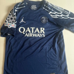 PSG x Jordan D. Doué - Snygg PSG matchtröja i mörkblått med vita detaljer, Jordan-logga och Qatar Airways-tryck på bröstet. Unika vingmönster på axlarna, Champions League-patch och UEFA Foundation-märke på ärmen. D. Doué och nummer 14 på ryggen. Tillverkad i lätt och ventilerande polyester. Kvaliten är hyfsat bra. Storlek L medn passar även M för en lösare fit