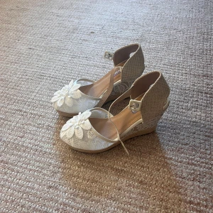 Vita kilklackade sandaler med spets - Snygga sandaler med kilklack från M. Adlina. Skorna är vita med spets och har en stor broderad blomma framtill. Baksidan är i beige textil och de har justerbart spänne vid ankeln. Perfekta för fester eller studenten.