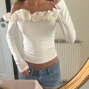 Vit offshoulder topp med volang - Superfin vit offshoulder topp med lång ärm och bred volang runt hela axelpartiet. Sitter tight och har en stilren, feminin look som passar perfekt till jeans eller kjol. Materialet känns mjukt och stretchigt.