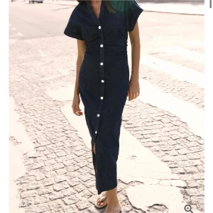 Marinblå jeansklänning med knappar - Snygg marinblå långklänning i jeansmaterial med korta ärmar och vita knappar hela vägen framtill. Klänningen har en slits nertill och markerad midja för en smickrande siluett. Använd några gånger skriv privat för flera bilder