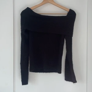 Svart offshoulder stickad tröja - Snygg svart stickad tröja med offshoulder-design och ribbad struktur. Tröjan har långa ärmar och en bred krage som vilar över axlarna för en trendig look. Perfekt att styla med jeans eller kjol för en cool vibe.