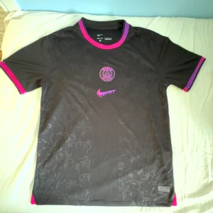 PSG x Nike svart fotbollströja - Svart PSG fotbollströja från Nike med diskret blommönster i tyget. Rosa och lila detaljer vid krage och ärmslut, samt rosa PSG-logga och Nike swoosh på bröstet. Tillverkad i lätt och ventilerande material, perfekt för träning eller match.