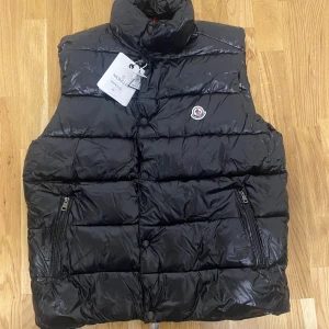 Svart dunväst från Moncler - Snygg svart dunväst från Moncler med hög krage och klassisk logga på bröstet. Västen har två dragkedjefickor framtill och stängs med både dragkedja och knappar. Perfekt för lager-på-lager och riktigt stilren look.
