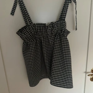 Rutig svartvit linnetopp med knytband - Supersöt linnetopp i svartvitt rutigt mönster med breda axelband som knyts upptill. Toppen har volangkant upptill. 