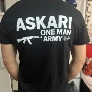 Svart ASKARI t-shirt med tryck - Svart t-shirt med vit text och motiv på både bröstet och ryggen. Stort tryck på ryggen med texten 'ASKARI ONE MAN ARMY' och en gevärsgrafik. Klassisk rund hals och korta ärmar. Skön passform, perfekt för dig som gillar streetwear.