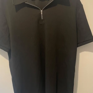 Svart ribbad pikétröja med half zip - Svart pikétröja från Manfinity (Shein) i storlek L. Tröjan har korta ärmar, ribbad struktur och en snygg silverfärgad half zip-dragkedja vid kragen. Perfekt för en clean och stilren look, funkar lika bra till jeans som till chinos.