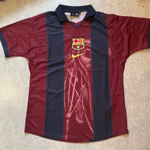 FC Barcelona x Cactus Jack fotbollströja - Snygg FC Barcelona fotbollströja i samarbete med Cactus Jack. Tröjan är röd och marinblå med breda vertikala ränder, klubbmärke och Nike-logga på bröstet. Baksidan har tryck med nummer 2, 'BARÇA' och Cactus Jack-grafik. Klassisk krage och korta ärmar. Tillverkad i polyester.