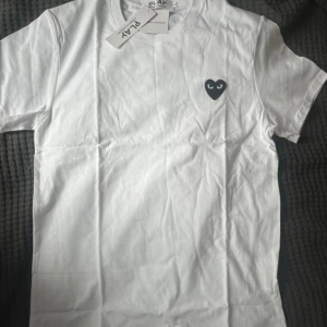 Vit t-shirt Comme des Garçons Play - Snygg vit t-shirt från Comme des Garçons Play med det ikoniska svarta hjärta-trycket på bröstet. Klassisk passform, rund hals och korta ärmar. Tillverkad i mjuk bomull, perfekt för en clean och stilren look. Står L passar S