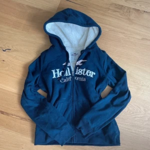 Hollister zip up hoodie  - Mysig Zip up Hoodie från Hollister med vit fluffig insida och huva. Hoodien har dragkedja framtill, snörning i huvan och stora fickor. Perfekt för kyliga dagar när du vill vara både varm och snygg.