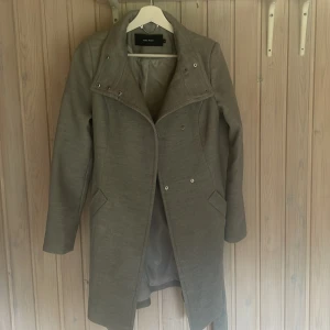 Grå kappa från Vero Moda XS - Snygg grå kappa från Vero Moda i storlek XS. Klassisk modell med bred krage, knappar och bälte i midjan. Fodrad insida och två fickor framtill. Perfekt för dig som gillar stilrena och enkla plagg med en modern touch.