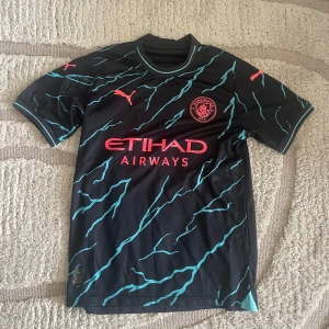 Manchester City Foden 47 Puma tröja - Manchester Citys officiella bortatröja från Puma med Foden 47 på ryggen. Svart bas med turkosa blixtmönster och röda detaljer, inklusive klubbmärke och Puma-logga. Tillverkad i lätt och ventilerande polyester, perfekt för vardagen eller träning.                       (Ej äkta city tröja)