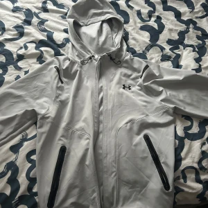 Ljusgrå vindjacka från Under Armour - Snygg ljusgrå vindjacka från Under Armour med huva och svarta detaljer. Jackan har dragkedja framtill, två fickor med svarta dragkedjor och justerbar huva. Perfekt för blåsiga dagar och sportiga outfits.