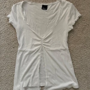 Gina tricot spets topp - Supersöt vit topp från Gina Tricot med v-ringning och spetsdetaljer längs halsringningen och framtill. Toppen har korta ärmar, snörningseffekt framtill och en liten slits. Perfekt för dig som gillar romantiska vibes och vill ha något extra i garderoben.