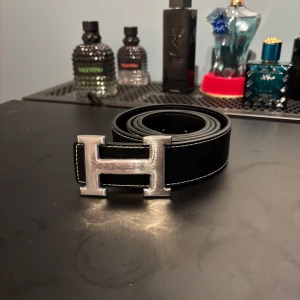H bälte - Säljer ett svart skärp med ikoniskt H-spänne i silverfärgad metall. Bältet är gjort av äkta läder och har vita sömmar som värkligen lyfter designen 🤩. Bältet är 110 cm långt och ettan de hetaste inom bälten. Hör av er vid frågor🥂🍾
