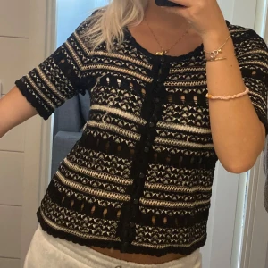 Mönstrad kofta från Indiska, svart/vit - Kortärmad kofta från Indiska i svart och vitt med snyggt stickat mönster. Koftan har knappar framtill, rund halsringning och vågiga kanter vid ärmslut och nederkant. Perfekt för dig som gillar unika detaljer och boho-vibe.