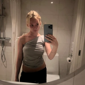 Grå oneshoulder topp med rynk - Säljer en snygg grå oneshoulder topp med rynkade detaljer längs sidan. Toppen är tight och har en cool asymmetrisk axelbandslös design som ger en trendig vibe. Perfekt att styla med jeans eller kjol för en modern look.