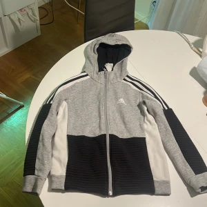 Grå och svart hoodie från Adidas - Säljer en snygg hoodie från Adidas i grått, svart och vitt. Tröjan har dragkedja framtill, huva och klassiska Adidas-ränder på axlarna. Materialet är mjukt och bekvämt, med ribbade svarta partier på ärmar och nederdel. Perfekt för en sportig look.