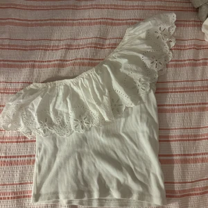 Vit offshoulder topp från Zara - Superfin vit offshoulder topp från Zara med bred volangkant och broderade blommor. Toppen är ärmlös och har en snygg, asymmetrisk design med spetsdetaljer längs volangen. Perfekt för en somrig look!
