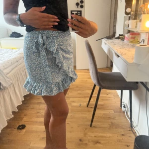 Blå blommig volangkjol med knyt - Supersöt blå kortkjol med vitt blommönster och volangkant. Kjolen har omlottmodell med knyt i midjan och extra volangdetalj framtill. Perfekt för sommarens alla dagar och ger en fräsch och trendig look.