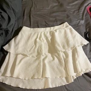 Vit volangkjol med struktur - Superfin vit kortkjol med flera lager volanger och snygg struktur i tyget. Kjolen har en lätt A-linjeform och resår i midjan för skön passform. Perfekt för dig som gillar en romantisk och trendig look. Andvänd ca 1 gång