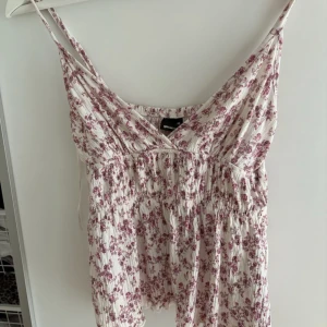 Blommig topp från Gina Tricot XS - Supersöt topp från Gina Tricot i XS med smala axelband och v-ringning. Vit botten med små rosa blommor, smockad midja och lätt, luftig känsla. Perfekt för varma dagar och ger en romantisk vibe. Har endast använt 1 gång och säljer för att det va i fel storlek! 🤗❤️