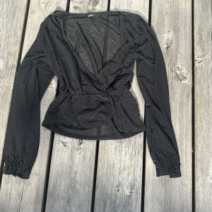 Svart prickig omlottblus från ONLY - Säljer en svart transparent omlottblus från ONLY med diskreta prickar i tyget. Blusen har långa ärmar med resår vid ärmslut och markerad midja. 