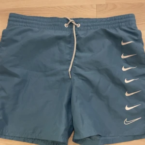 Blå Nike shorts med snörning XS - Snygga blå bad/tränings shorts från Nike i storlek XS. De har elastisk midja med vit snörning och flera vita Nike-loggor på ena benet. Perfekta för stranden eller träning, tillverkade i lätt och snabbtorkande material.