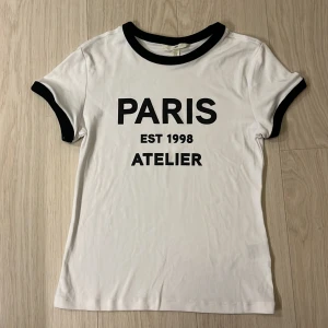Vit t-shirt H&M  - Vit t-shirt från H&M med svart text 'PARIS EST 1998 ATELIER' på bröstet. Svarta ribbade kanter vid halsringning och ärmslut ger en snygg kontrast. Klassisk passform och tillverkad i mjuk bomull. Tröjan är knappt använd. 