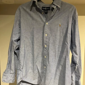Blå skjorta från Ralph Lauren - Klassisk blå skjorta från Ralph Lauren i 100% bomull. Skjortan har lång ärm, knappar framtill och den ikoniska broderade loggan på bröstet. Perfekt för en clean och stilren look. Skjortan är i bra skick utan några defekter! Storleken är 8, vilket ungefär motsvarar L. Bara att höra av sig vid eventuella frågor!😉