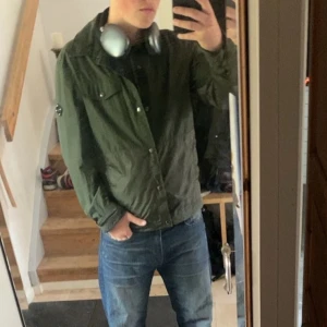 Grön vindjacka med knappar - En Snygg grön vindjacka från j.lindberg med klassisk krage och knäppning framtill. Jackan har en avslappnad passform och är perfekt för dig som gillar en enkel men stilren look. Passar bra till jeans och sneakers för en casual vibe. Är S men passar M