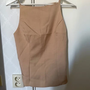 Beige topp med knyt  - Stilren beige topp med smala, justerbara axelband och omlottdetalj i ryggen som knyts i sidan. Toppen har rak halsringning och är ärmlös, perfekt för en trendig och minimalistisk look.