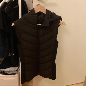 Svart dunväst med huva - Säljer en svart dunväst med huva och dragkedja framtill. Västen har en quiltad design med snygga diagonala sömmar och är lite längre i modellen. Perfekt för lager-på-lager och chillare dagar när du vill hålla värmen utan att ha en jacka.