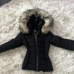 Svart pufferjacka med fuskpäls - Svart pufferjacka från Marikoo med stor huva kantad av fuskpäls. Jackan har quiltad design, dragkedja framtill och två fickor med dragkedja. Figursydd modell som ger en snygg siluett och håller dig varm under kalla dagar.